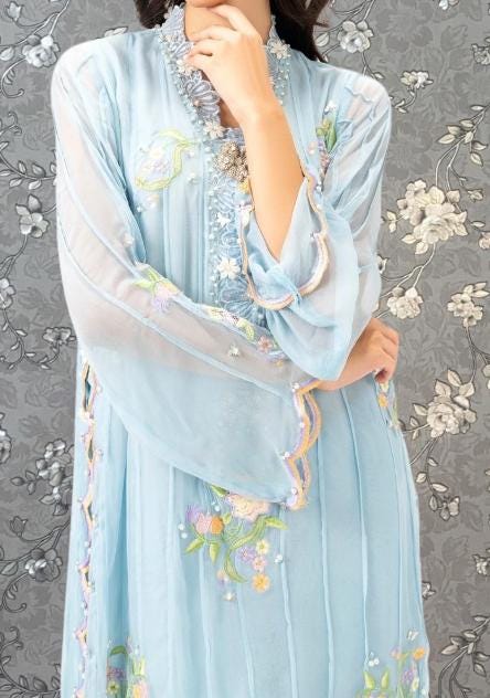A woman wearing a light blue dress with intricate floral embroidery stands against a gray floral background.