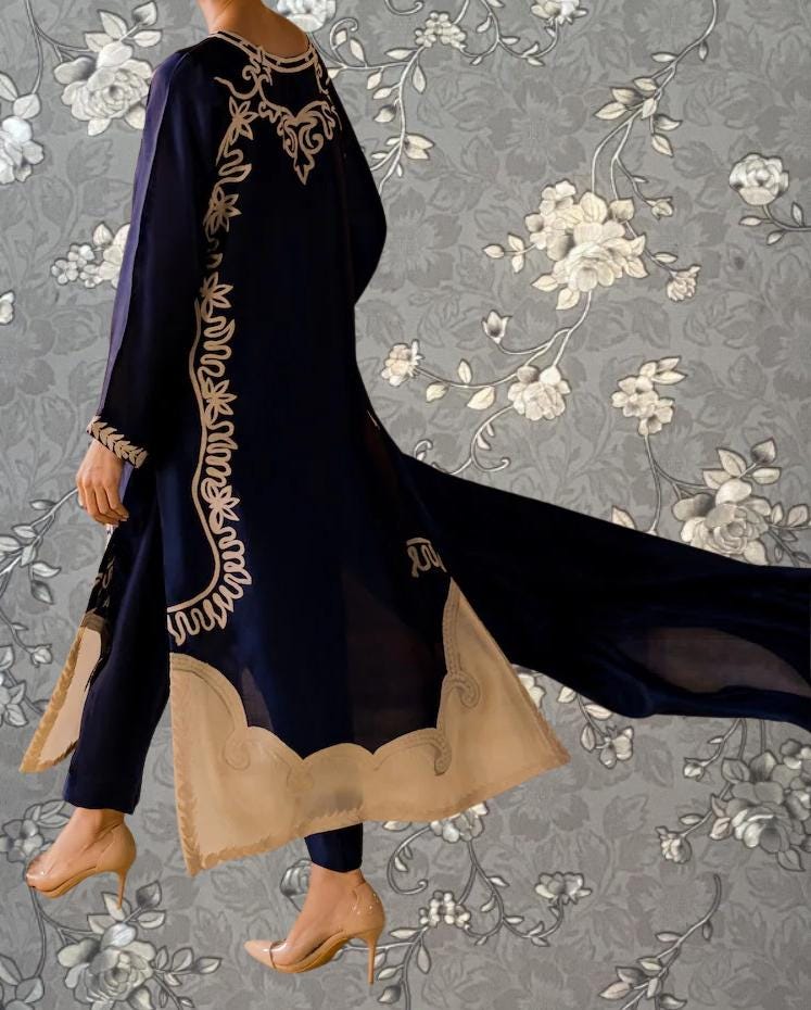 A person wearing a long, dark blue dress with gold embroidered patterns on the sleeves and hem, standing against a gray floral background.