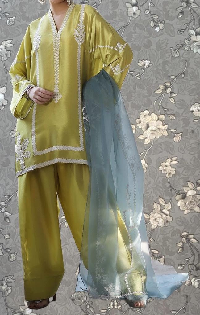 A person wearing a yellow and blue outfit with intricate embroidery, standing against a gray floral background.