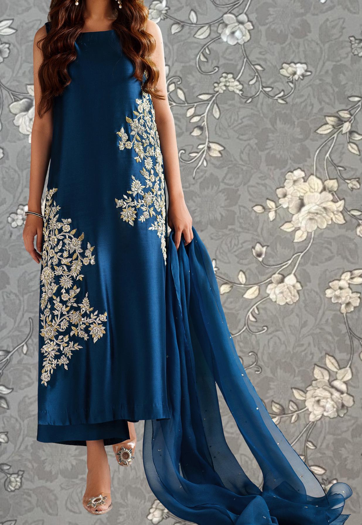 A woman wearing a long blue dress with white floral embroidery stands against a gray floral background.