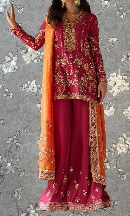 A woman wearing a vibrant red and orange traditional Indian outfit with intricate embroidery and patterns, standing against a gray floral background.