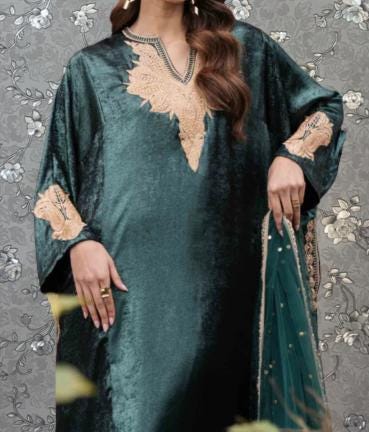 A woman wearing a long, green velvet dress with gold embroidery stands against a gray floral background.