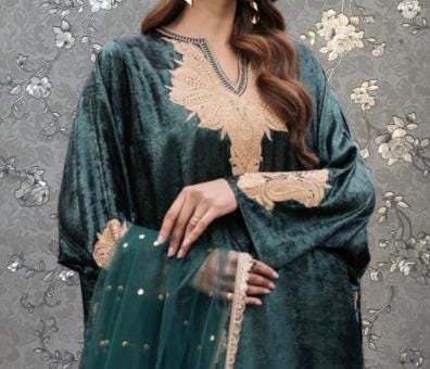A woman wearing a green velvet dress with gold embroidery and a matching green shawl stands against a gray floral background.