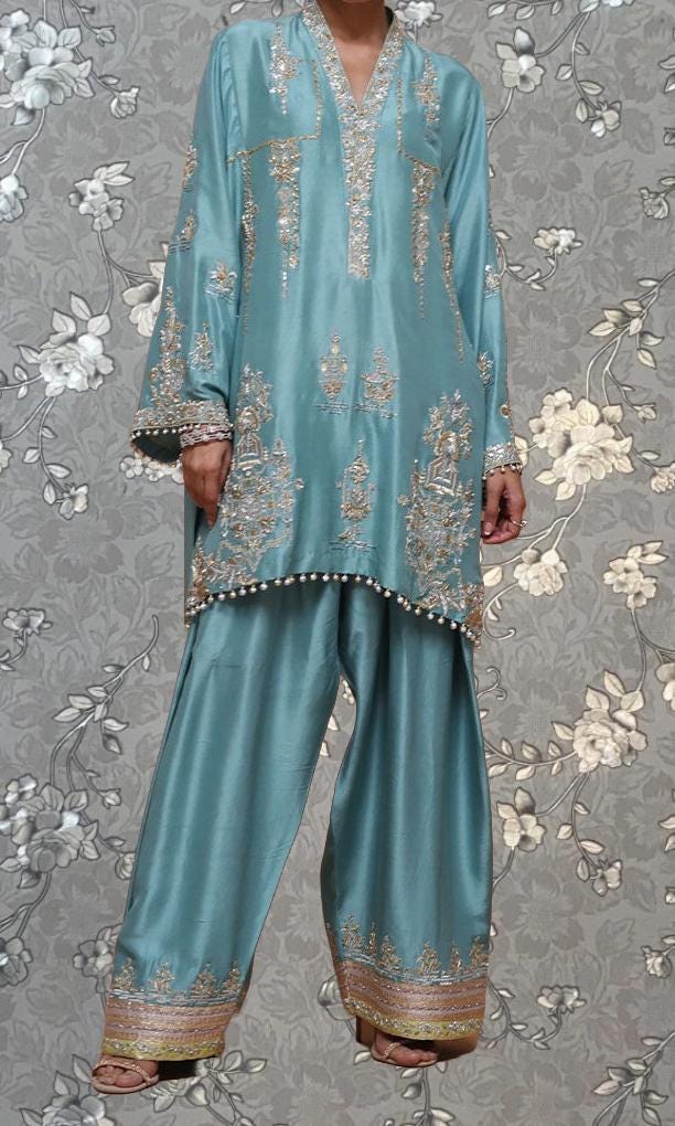 A person wearing a turquoise and gold embroidered dress stands against a gray floral background.