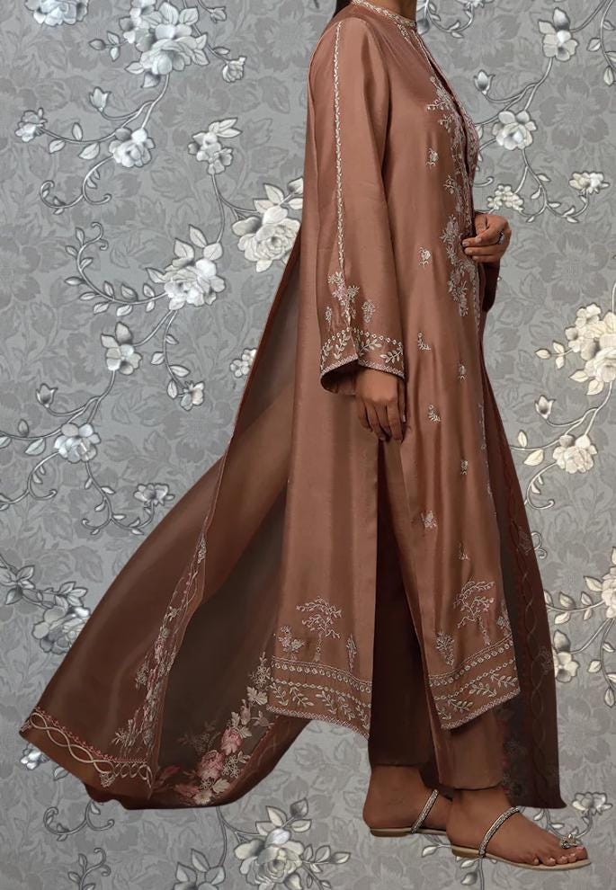 A woman wearing a long brown dress with intricate embroidery stands against a gray floral background.