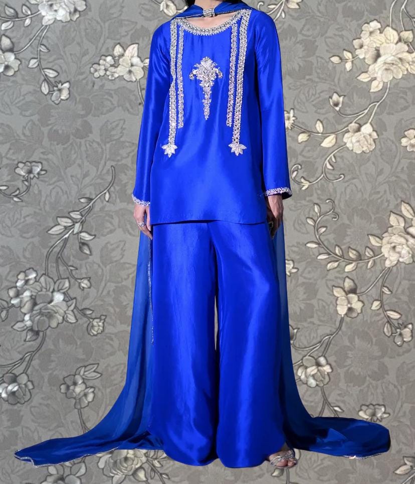 A person wearing a long, blue dress with intricate embroidery and a matching blue skirt, standing against a gray floral background.