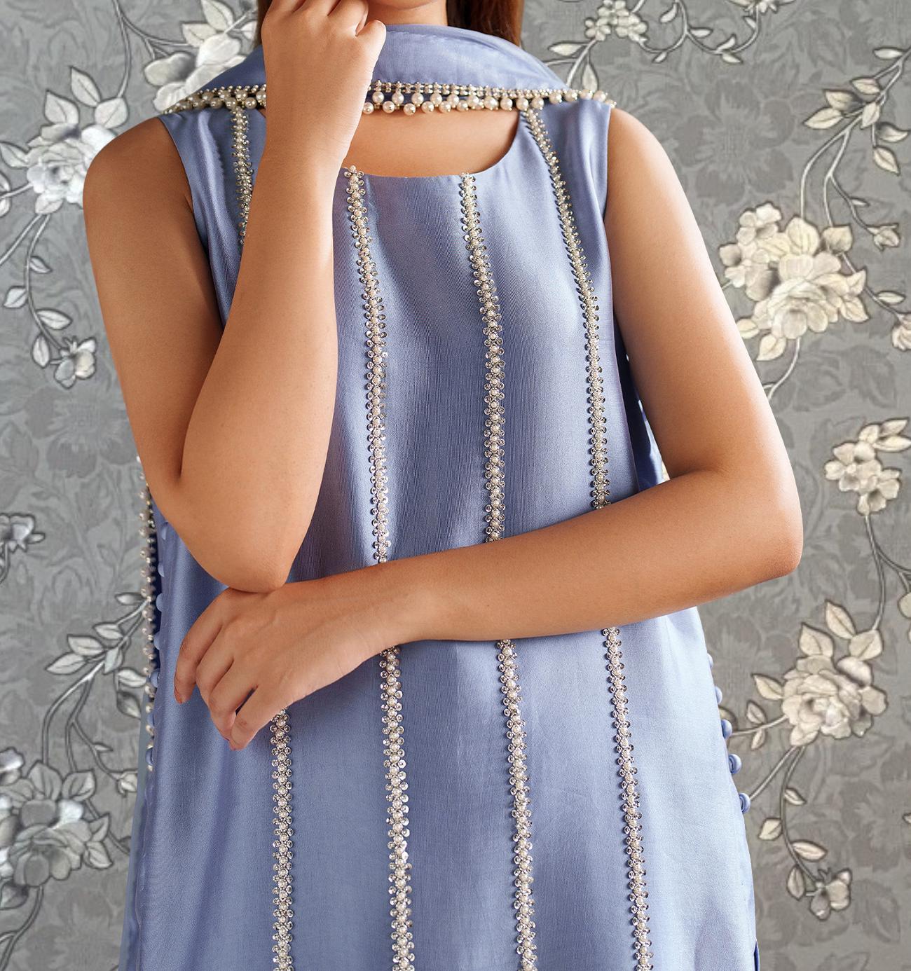 A woman wearing a sleeveless blue dress with a floral pattern and beaded details, posing with her hand on her chin.