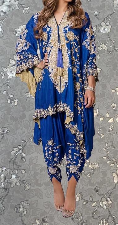 A woman wearing a blue and gold embroidered dress with a floral pattern, standing against a gray floral background.