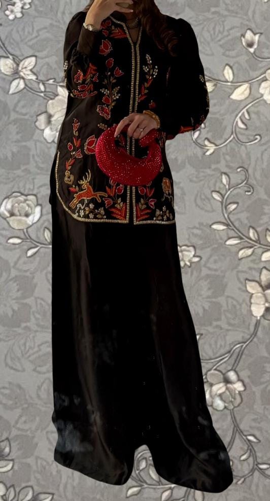 A woman wearing a long black dress with a floral pattern and a red clutch purse is standing against a gray floral background.