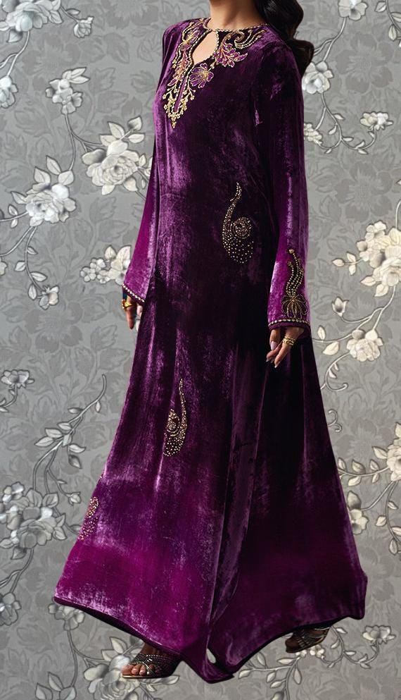 A woman wearing a long, purple velvet dress with intricate gold embroidery stands against a gray floral background.