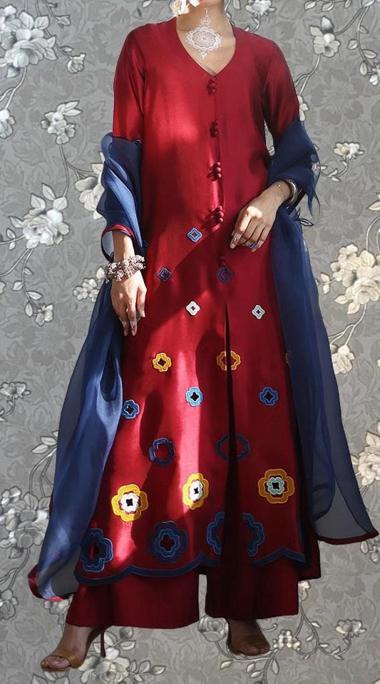 A person wearing a red dress with blue and yellow floral embroidery, standing against a gray floral background.