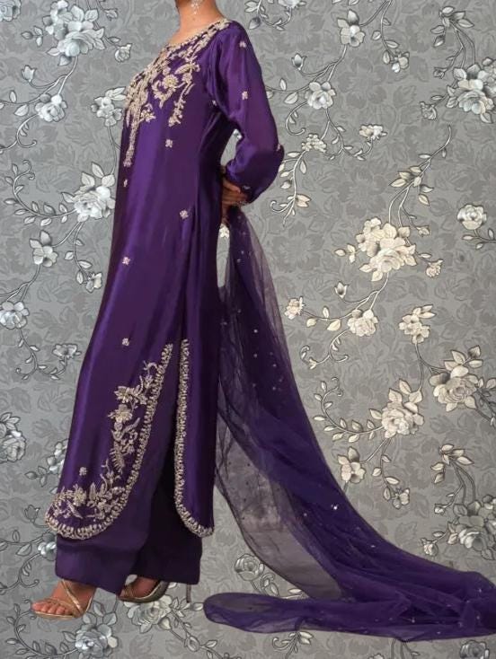 A woman wearing a long purple dress with intricate embroidery stands against a gray floral background.