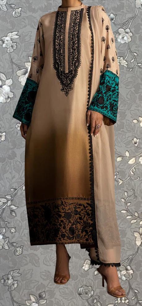 A person wearing a long, beige dress with intricate black embroidery and teal accents, standing against a gray floral background.