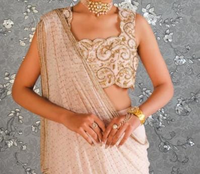 A woman wearing a white and gold saree with a gold and beige blouse, accessorized with jewelry, posing against a gray floral background.
