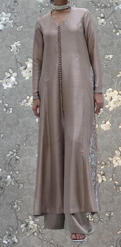 A person wearing a long, beige dress with a floral pattern on the sleeves and hem, standing against a gray floral background.