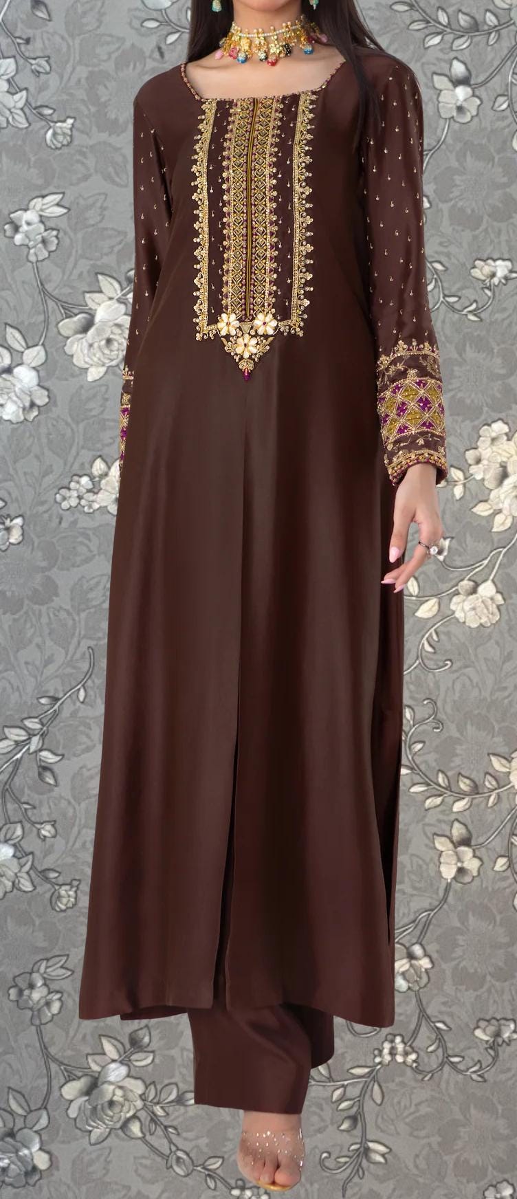 A woman wearing a brown dress with intricate embroidery and gold accents, standing against a gray floral background.