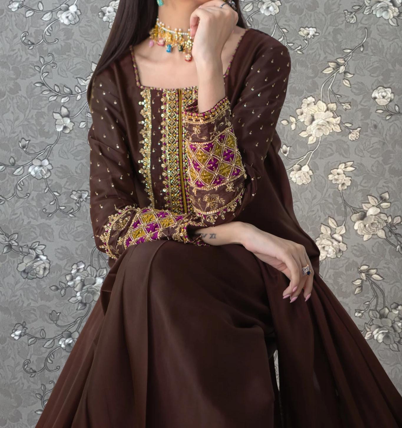 A woman wearing a brown dress with intricate embroidery and jewelry poses against a floral background.