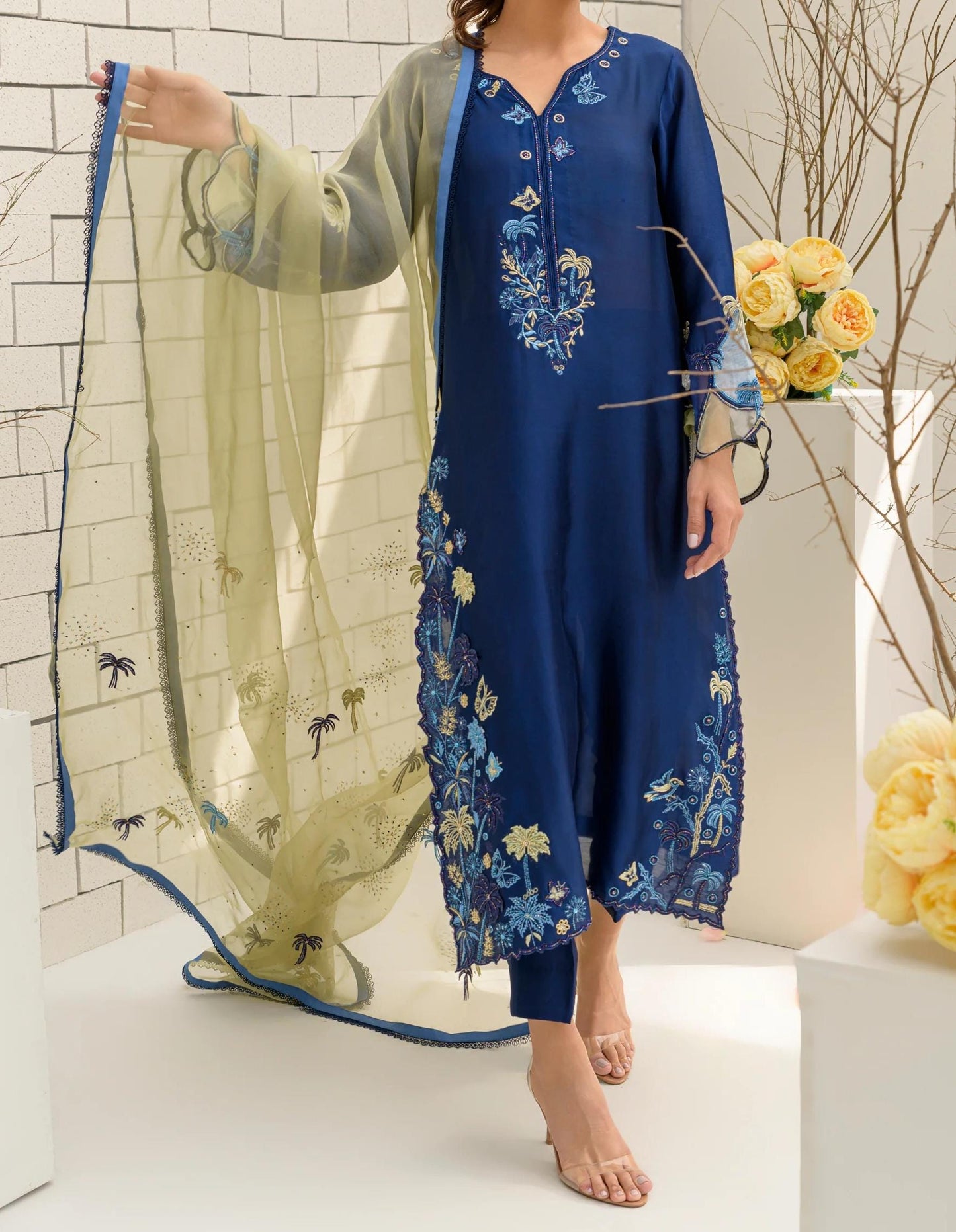 A woman wearing a blue dress with intricate embroidery stands in front of a white brick wall, holding a bouquet of yellow flowers.