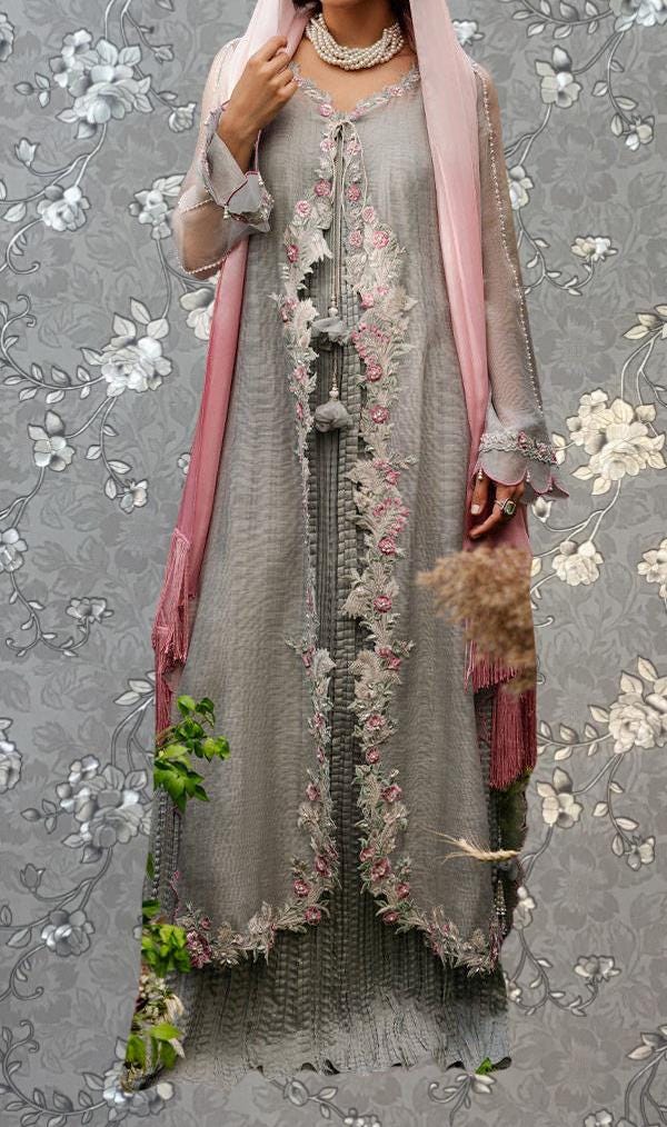 A woman wearing a long, gray dress with intricate embroidery and a pink shawl, standing against a gray floral background.
