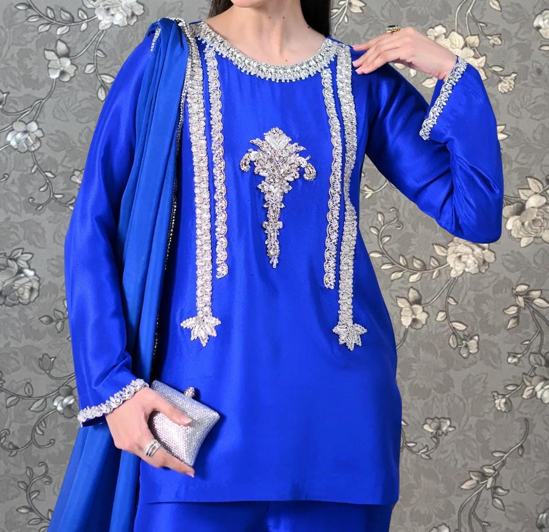 A woman wearing a blue dress with intricate silver embroidery and a matching blue shawl stands against a gray floral background.