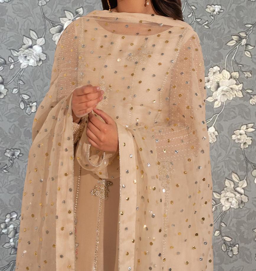 A woman wearing a beige dress with a sheer, polka-dotted pattern, standing against a gray floral background.