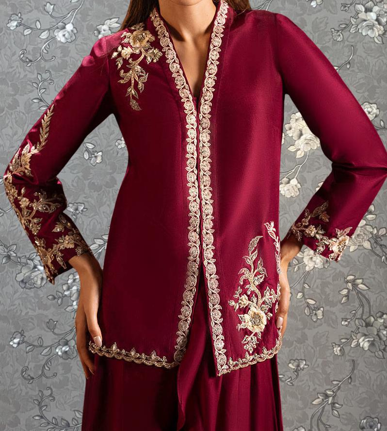 A woman wearing a red and gold embroidered dress with a high collar and long sleeves, posing with her hands on her hips against a gray floral background.