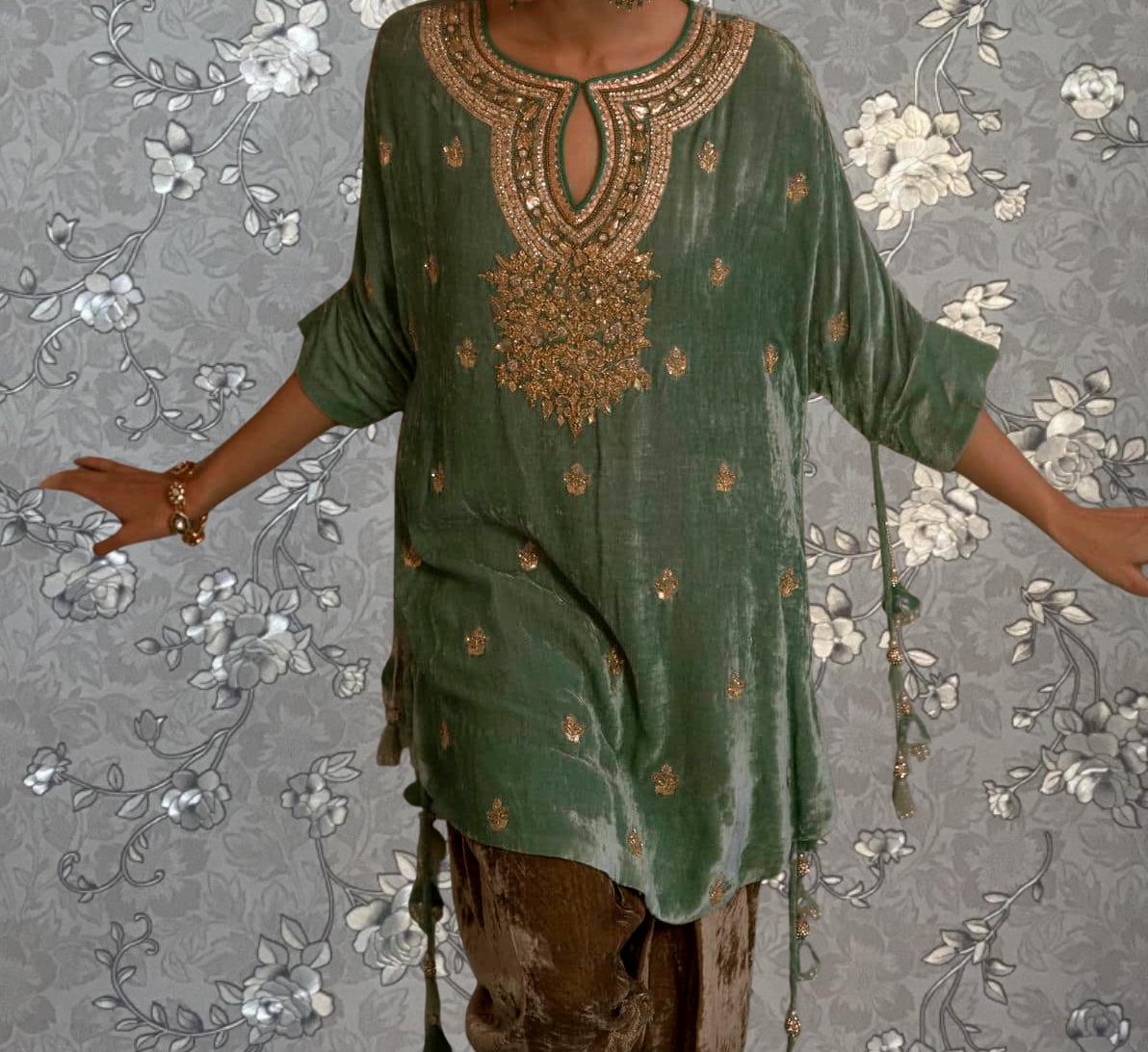A person wearing a green velvet dress with gold embroidery stands in front of a gray floral wallpaper.