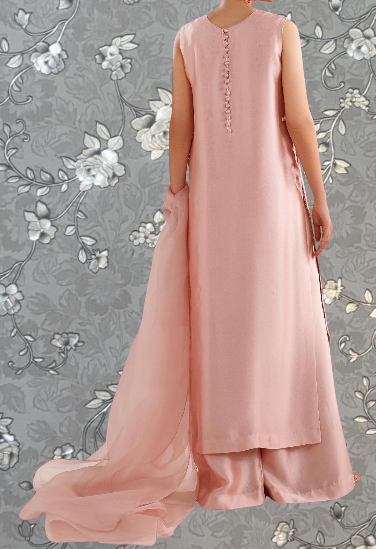 A woman wearing a long, sleeveless pink dress with a flowing skirt, standing against a gray floral background.