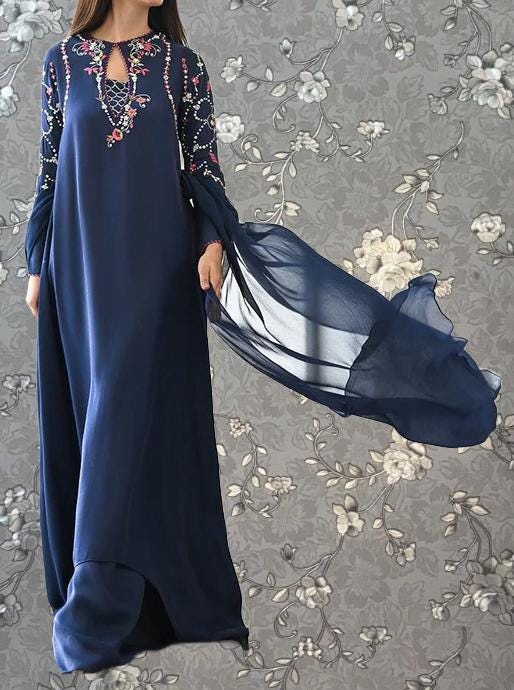 A woman wearing a long navy blue dress with floral embroidery and a flowing blue scarf is standing against a gray floral background.