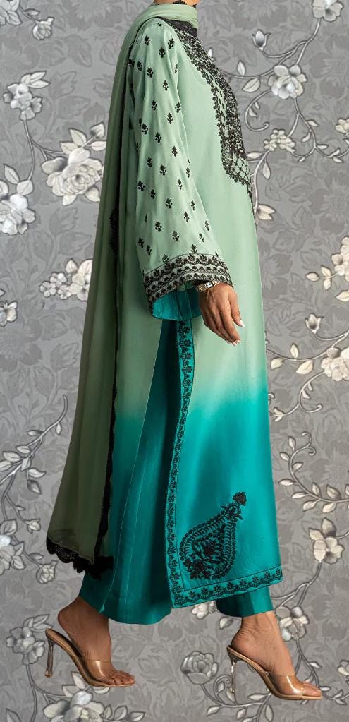 A person wearing a long, green and teal dress with intricate embroidery and floral patterns, standing against a gray floral background.