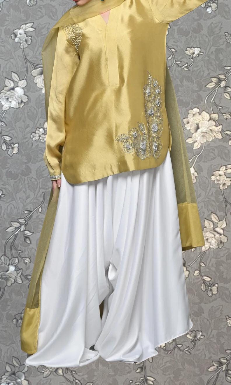 A gold-colored dress with a floral embroidered design on the back, paired with a white skirt, is shown against a gray floral background.