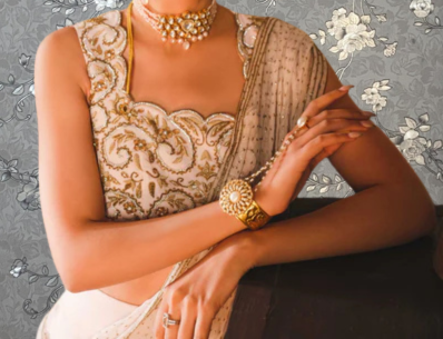 A woman wearing a gold and white dress with intricate embroidery and jewelry poses elegantly against a gray floral background.