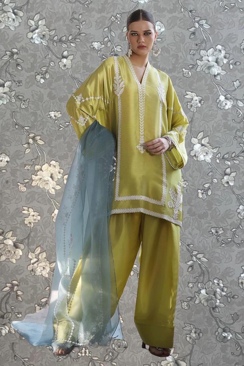 A woman wearing a yellow and white outfit with intricate embroidery stands against a gray floral background.