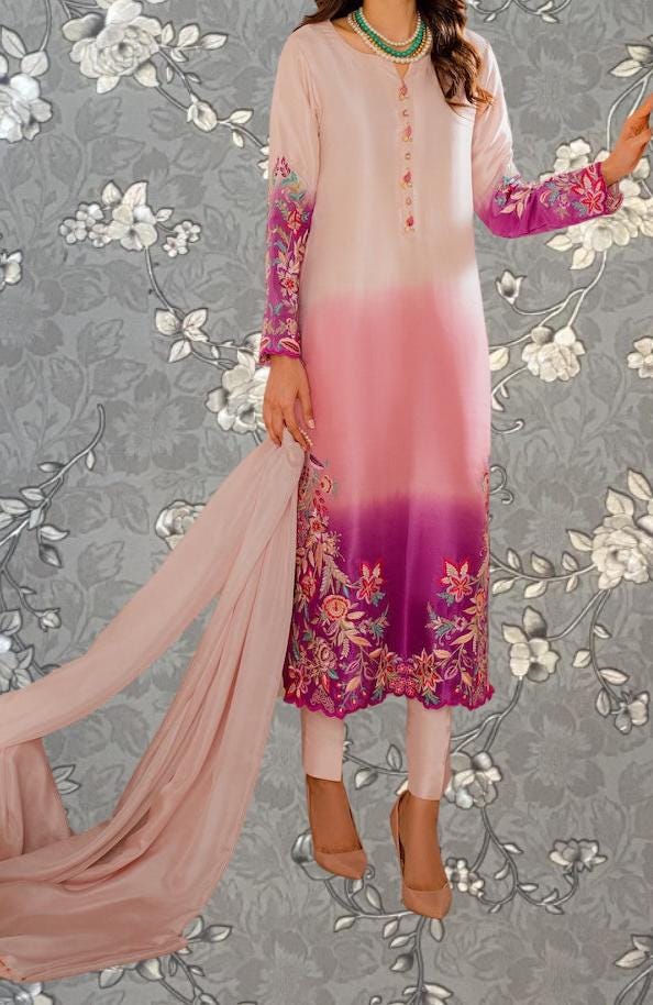 A woman wearing a pink and purple dress with floral embroidery, standing against a gray floral background.