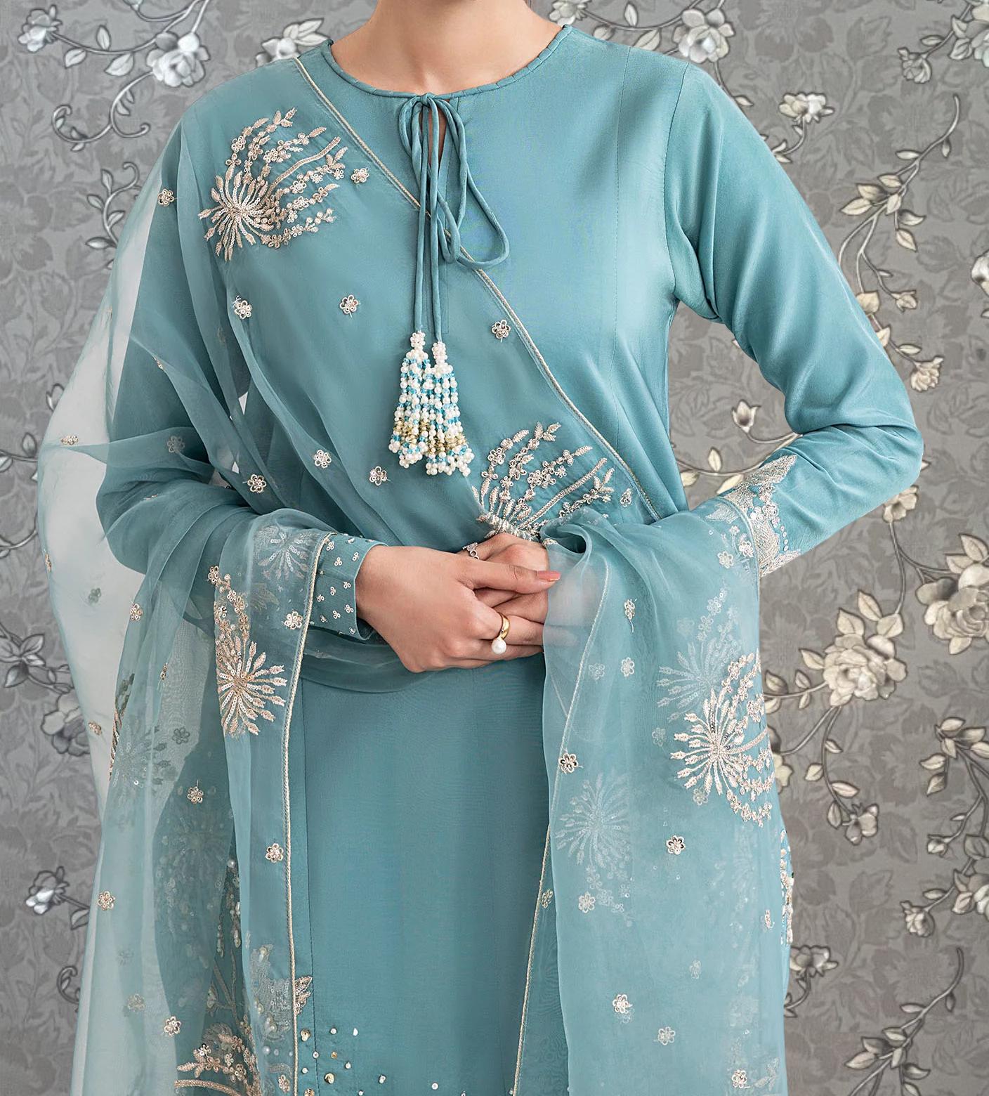 A person wearing a teal-colored dress with intricate embroidered designs, including flowers and leaves, stands against a gray floral background.