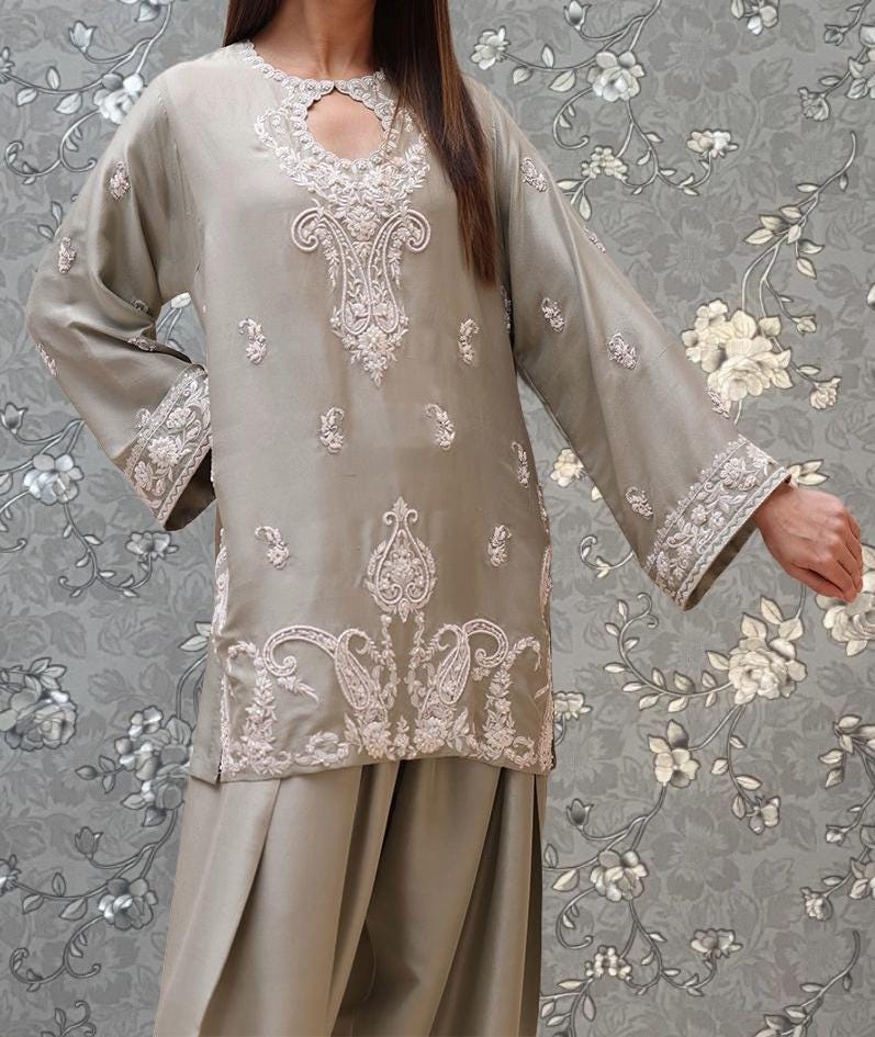 A woman wearing a grey dress with intricate embroidered patterns on the front and sleeves, standing against a gray floral background.
