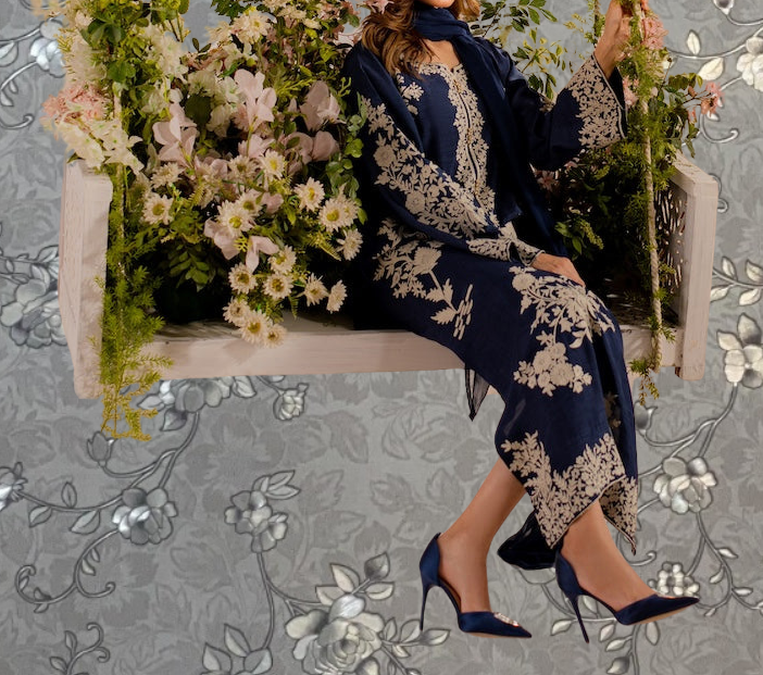 A woman in a blue and gold dress sits on a floral-patterned bench, surrounded by a variety of flowers and greenery.