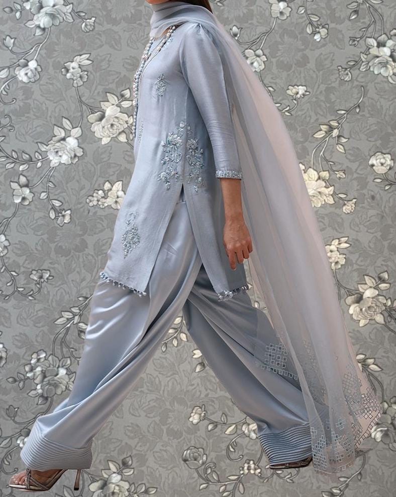 A person wearing a gray dress with intricate embroidery and a long, flowing gray shawl is walking against a gray floral background.