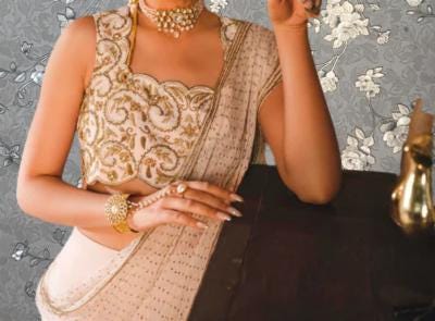 A woman wearing a gold and white saree with intricate embroidery and jewelry poses elegantly in front of a floral patterned background.