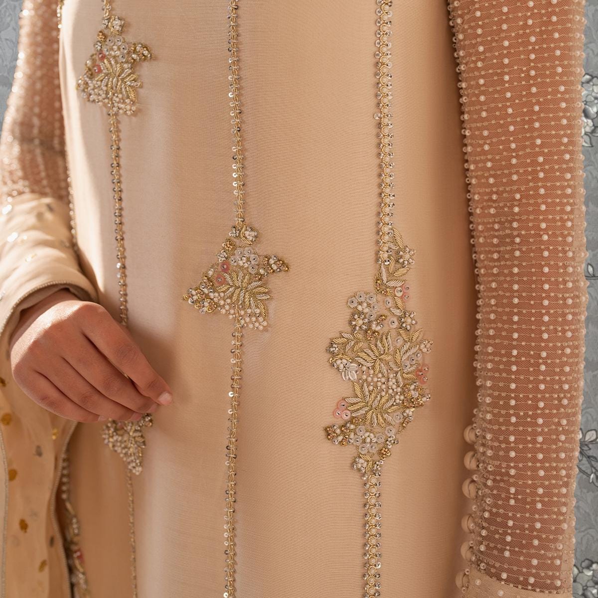 A close-up view of a person's hand wearing a beige dress with intricate gold embroidery and a beaded sleeve.