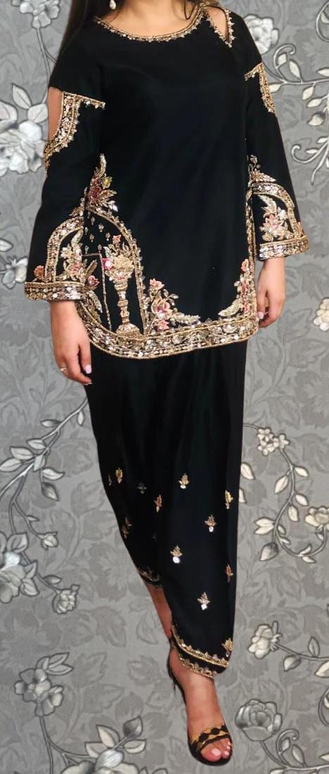 A woman wearing a black dress with gold embroidery and floral patterns, standing against a gray floral background.