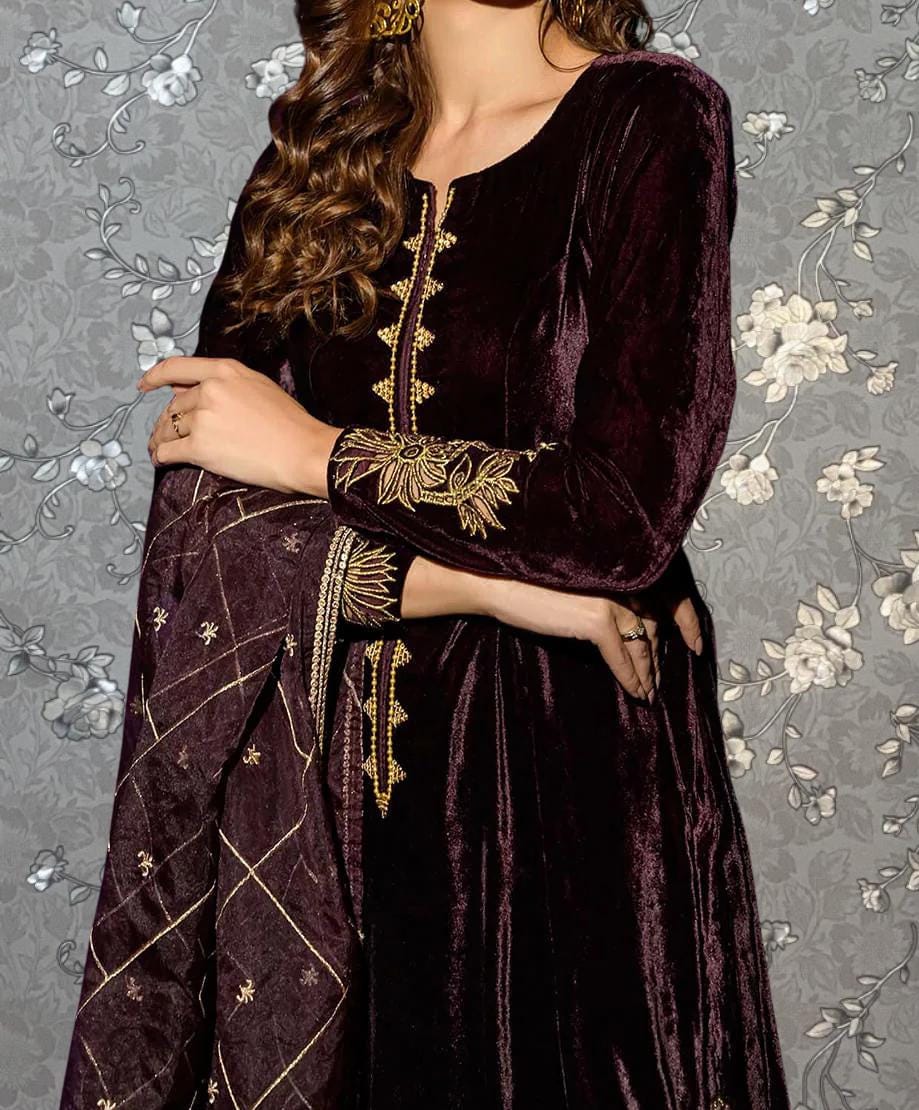 A woman wearing a long, burgundy velvet dress with gold embroidery stands against a gray floral background.