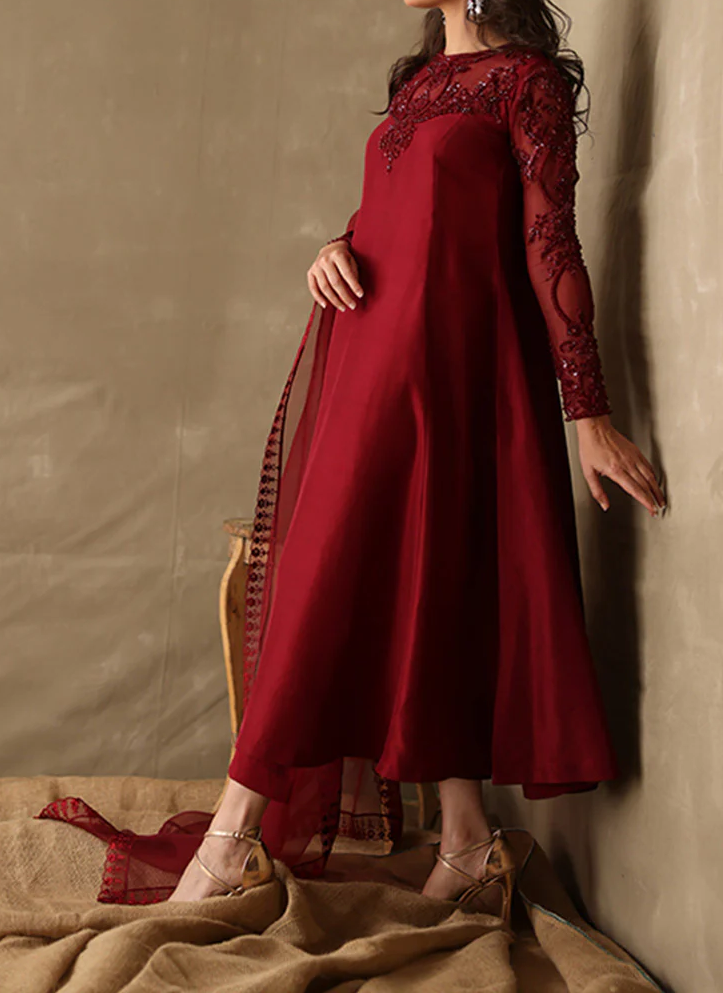 A woman wearing a long, burgundy dress with lace sleeves and a high neckline stands against a beige background.