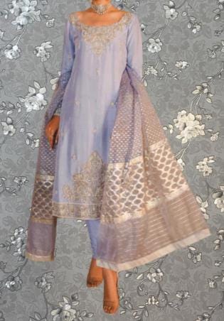 A woman wearing a lavender and gold patterned dress with a matching shawl, standing against a gray floral background.
