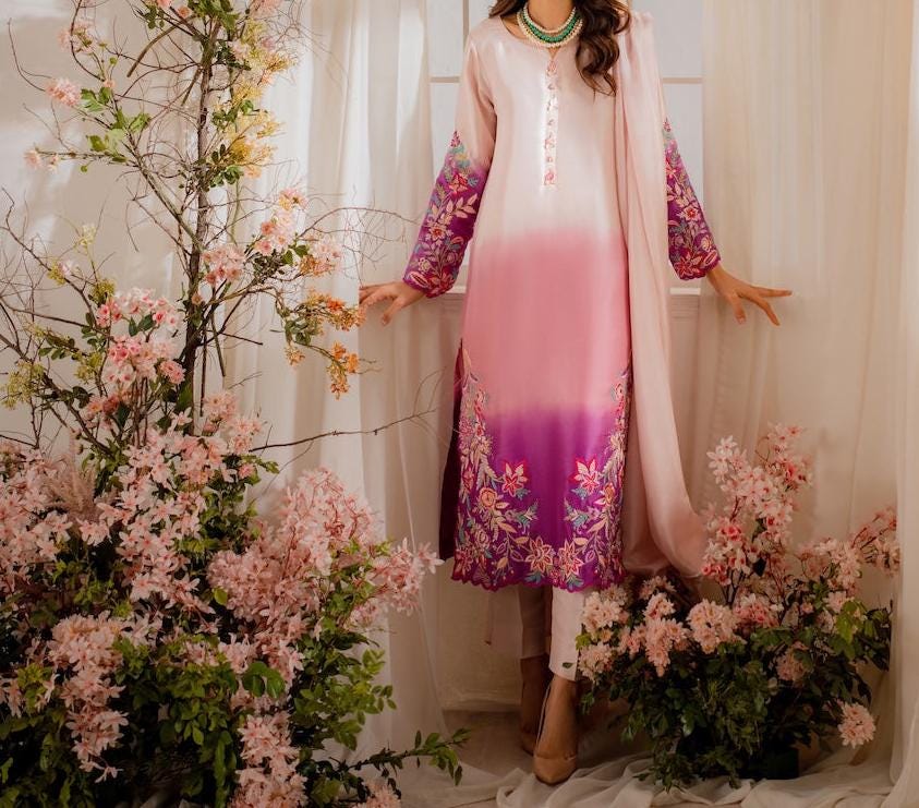 A woman in a pink and purple floral dress stands in front of a window, surrounded by pink and white flowers and a white curtain.