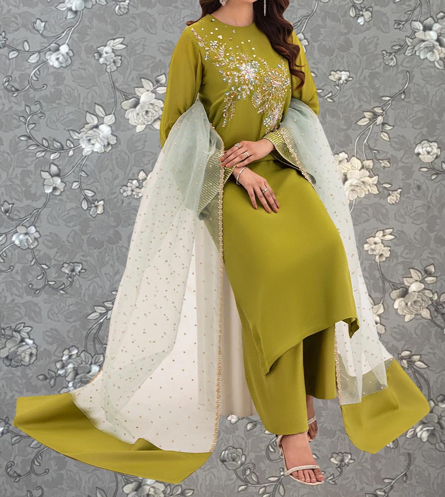 A woman wearing a green dress with gold embroidery and a white shawl, standing against a gray floral background.