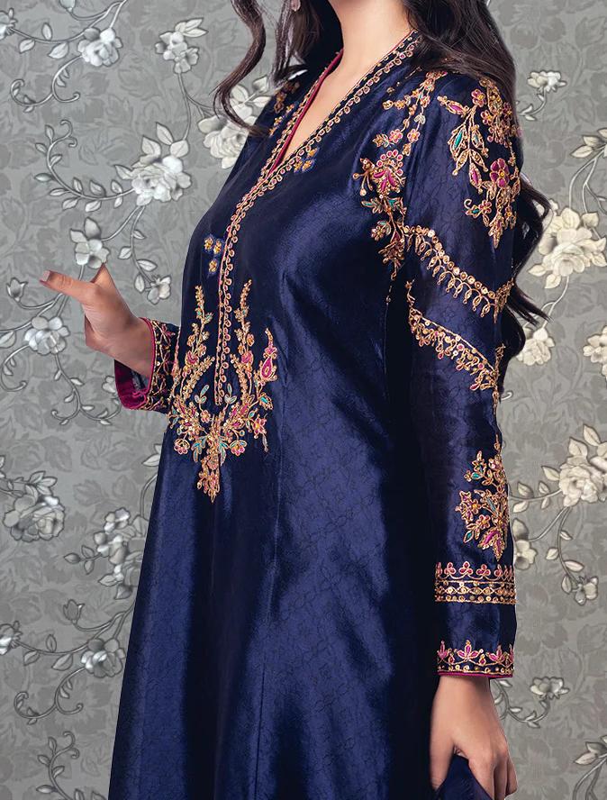 A woman wearing a blue dress with intricate gold embroidery stands against a gray floral background.