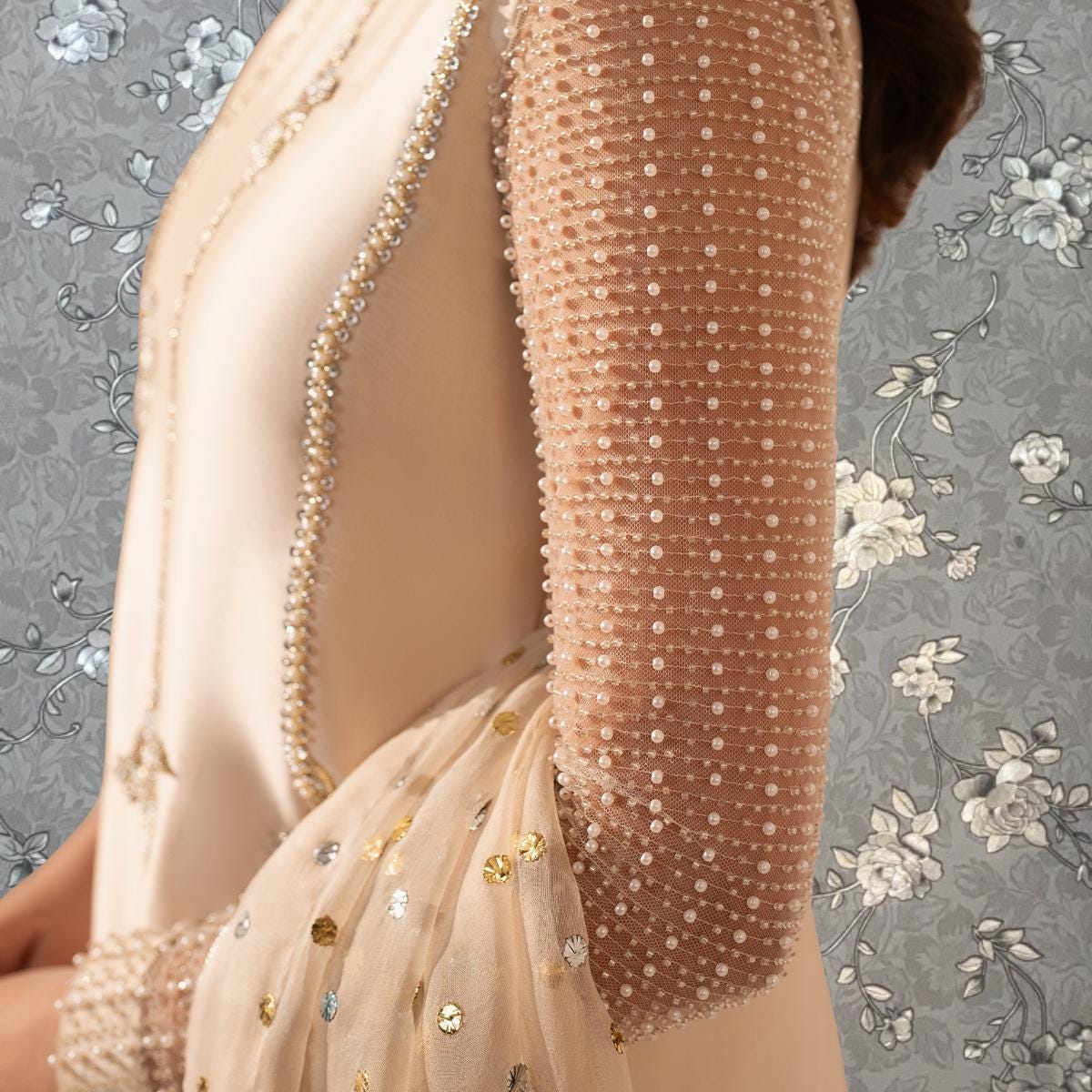 A close-up of a person wearing a beige dress with a sheer overlay and beaded details, standing against a floral patterned background.