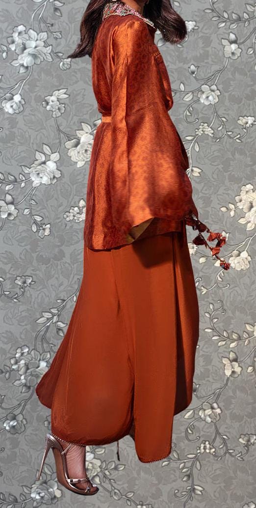 A woman wearing a long, flowing orange dress with a high collar and a matching orange blouse stands against a gray floral background.