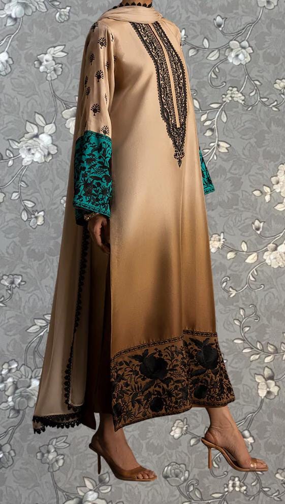 A person wearing a long, beige dress with intricate black and teal embroidery, standing against a gray floral background.